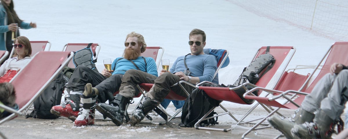 Still from Force Majeure. Two 30-something men are sitting in deck chairs in the snow, wearing ski boots, drinking a beer.